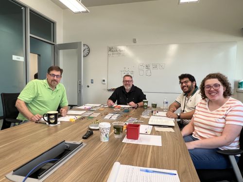 Cory Heald sits at a conference table with Merit Network staff during a game‑testing session. Game materials, notes, and tokens are spread across the table in a meeting room.